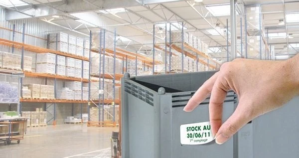 Retrait à la main d'une étiquette enlevable d'inventaire posée sur un bac plastique, dans un entrepôt logistique